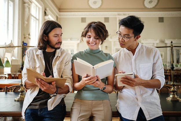 Grupo de jovens estudantes multinacionais casuais felizmente estudando com livro juntos na biblioteca da universidade