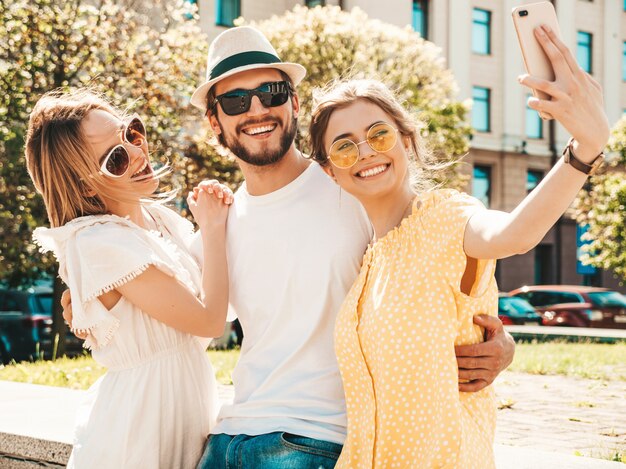 Grupo de jovens três amigos elegantes na rua. Homem e duas miúdas giras vestidas com roupas de verão casual. Modelos sorridentes se divertindo em óculos de sol. Mulheres e cara fazendo foto selfie em smartphone