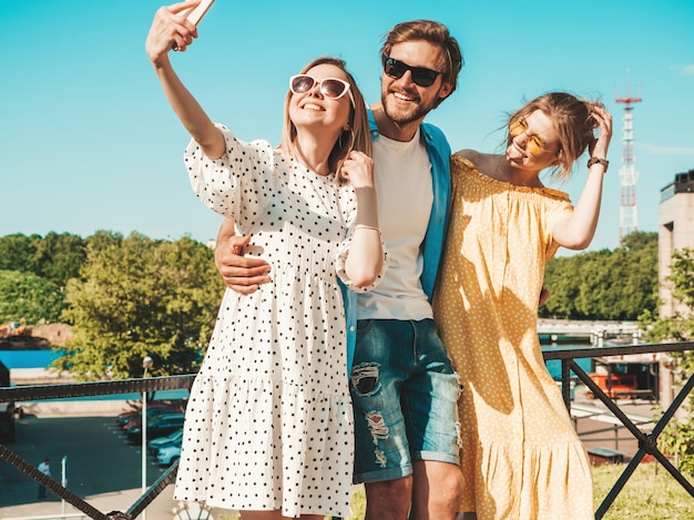 Grupo de jovens três amigos elegantes na rua. Homem e duas miúdas giras vestidas com roupas de verão casual. Modelos sorridentes se divertindo em óculos de sol. Mulheres e cara fazendo foto selfie em smartphone