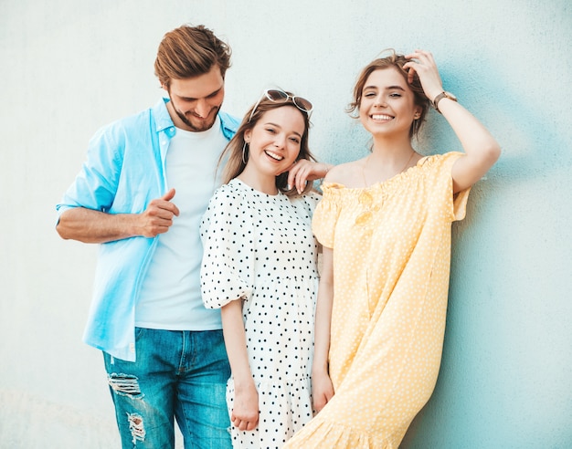 Grupo de jovens três amigos elegantes posando na rua. homem moda e duas miúdas giras vestidas com roupas de verão casual. modelos sorridentes se divertindo perto da parede. mulheres alegres e cara ao ar livre