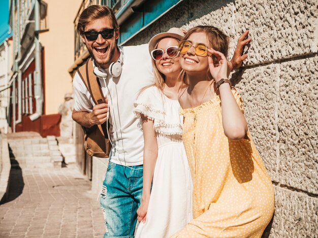 Grupo de jovens três amigos elegantes posando na rua. Homem moda e duas miúdas giras vestidas com roupas de verão casual. Sorrindo modelos se divertindo em óculos de sol. Mulheres alegres e cara ao ar livre