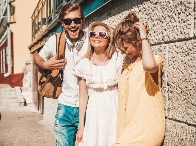 Grupo de jovens três amigos elegantes posando na rua. Homem moda e duas miúdas giras vestidas com roupas de verão casual. Sorrindo modelos se divertindo em óculos de sol. Mulheres alegres e cara ao ar livre