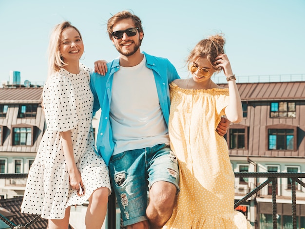 Grupo de jovens três amigos elegantes posando na rua. homem moda e duas miúdas giras vestidas com roupas de verão casual. sorrindo modelos se divertindo em óculos de sol. mulheres alegres e cara ao ar livre