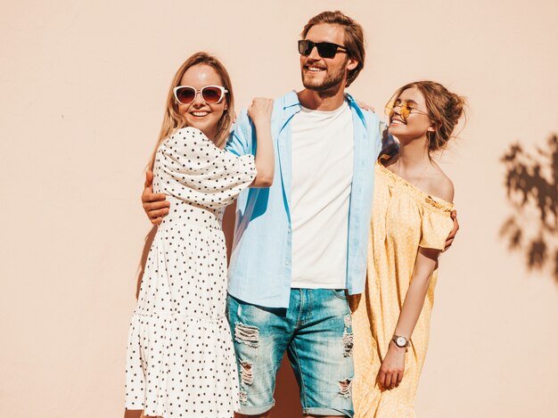 Grupo de jovens três amigos elegantes posando na rua. Homem moda e duas miúdas giras vestidas com roupas de verão casual. Sorrindo modelos se divertindo em óculos de sol. Mulheres alegres e cara enlouquecendo