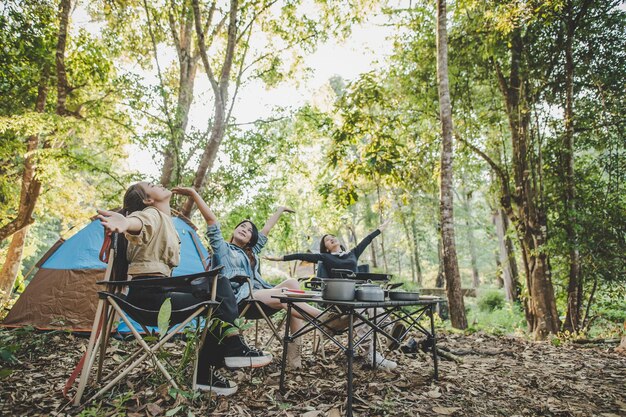 Grupo de mulheres bonitas sentadas na cadeira de acampamento na frente da barraca para relaxar enquanto acampam na floresta natural com felicidade juntos copiam o espaço