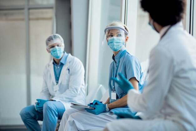 Grupo de profissionais de saúde com roupas de trabalho protetoras falando enquanto está sentado em um corredor na clínica médica