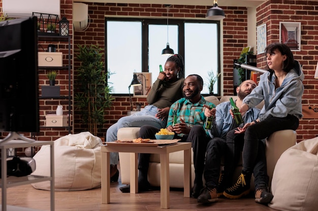 Grupo diversificado de amigos assistindo filme na televisão, comendo pipoca e bebendo garrafas de cerveja em uma reunião divertida. pessoas assistindo filme no programa do canal de tv, desfrutando de atividades de lazer.