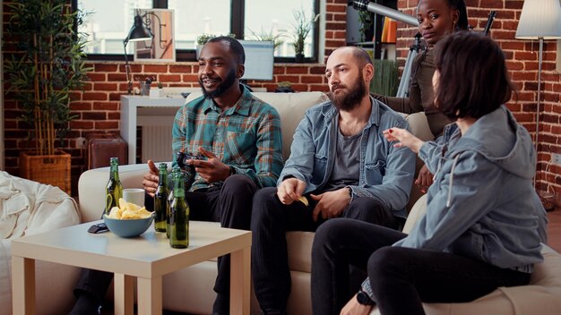 Grupo diversificado de amigos perdendo videogames no console, jogando competição online com controlador na reunião. Sentindo-se frustrado com o jogo perdido, bebendo cerveja e se divertindo.