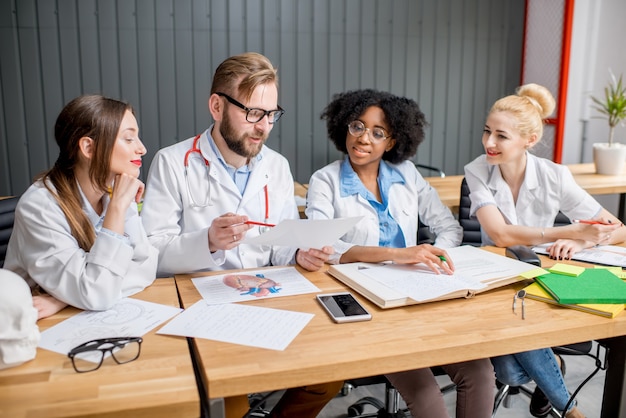 Grupo multiétnico de estudantes de medicina uniformizados discutindo sentados à mesa com diferentes materiais médicos na sala de aula