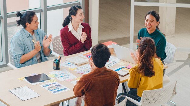 Grupo multirracial de jovens criativos da Ásia em smart casual wear, discutindo negócios, aplaudindo, rindo e sorrindo juntos em uma reunião de brainstorm no escritório. Conceito de sucesso do trabalho em equipe do colega de trabalho.