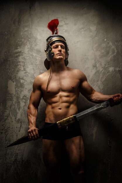 Guerreiro musculoso com espada e capacete posando em frente a parede de concreto