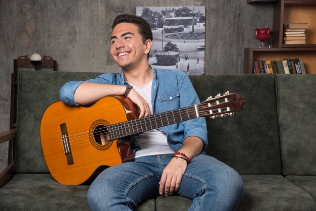 Guitarrista positivo segurando uma bela guitarra e sentado no sofá. Foto de alta qualidade