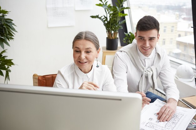 Habilidosa e sorridente engenheira-chefe madura usando o programa CAD no computador enquanto sua alegre jovem assistente estuda a planta na mesa na frente dele. Sucesso, trabalho em equipe e colaboração