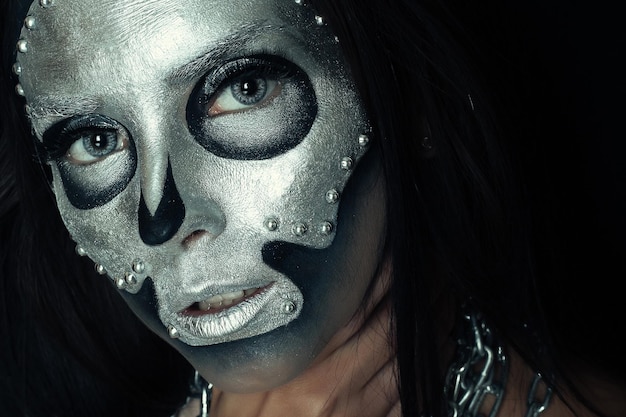 Halloween, feriados, estilo de vida, pessoas, beleza, conceito criativo - halloween e tema de maquiagem criativa: modelo de menina bonita com corpo preto com máscara de prata caveira pintada em fundo escuro no estúdio