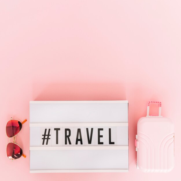 Hashtag com texto de viagens na lightbox com óculos escuros e bolsa de viagem em miniatura no fundo rosa