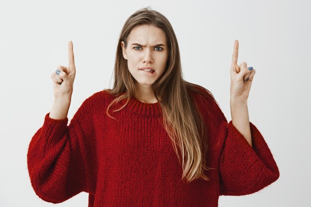 Hesitante, modelo feminino caucasiano atraente em um moderno suéter solto vermelho levantando os dedos indicadores e apontando para cima, mordendo o lábio, franzindo a testa, sendo intenso e preocupado, estando em dúvida e tomando decisões