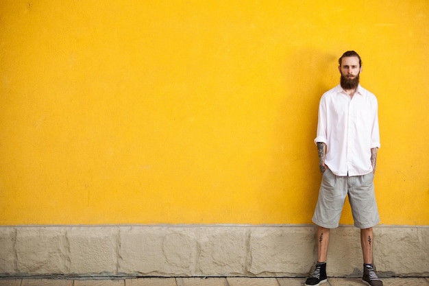 Hipster barbudo tatuado na parede amarela posando ao ar livre