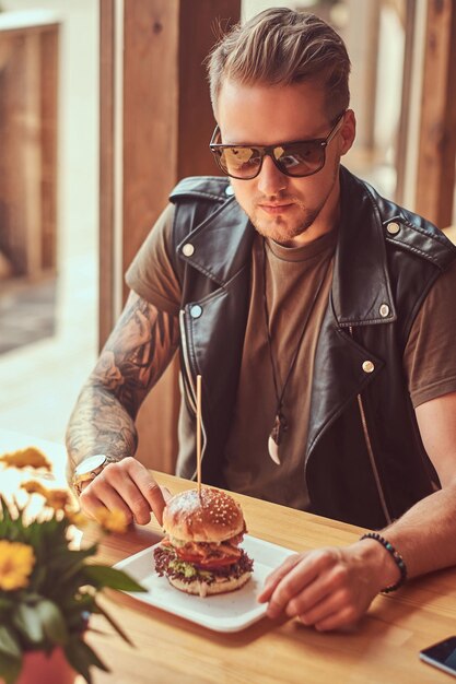 Hipster com um corte de cabelo elegante e barba se senta em uma mesa em um café na estrada, bebe um refrigerante em um dia quente.