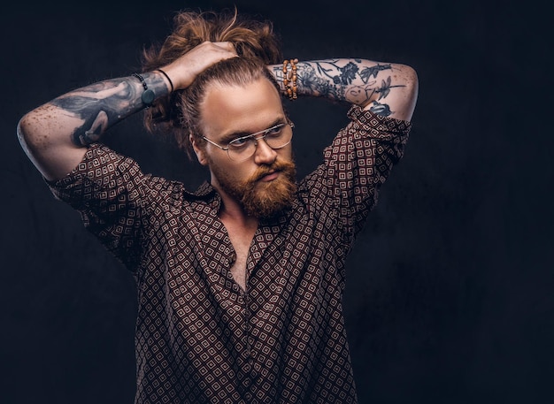 Foto grátis hipster de homem ruivo tatuado corrige seu cabelo exuberante vestido com uma camisa marrom, de pé em um estúdio. isolado no fundo escuro.
