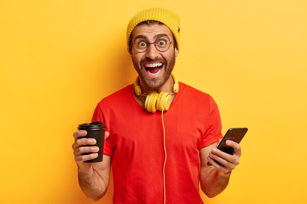 Hipster feliz cria novo perfil nas redes sociais, ri de felicidade, segura aparelho eletrônico moderno, bebe café