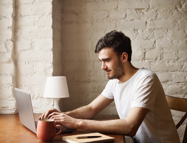 Hipster feliz em uma camiseta branca, sente-se embaixo da mesa e digite uma mensagem para uma amiga no caderno