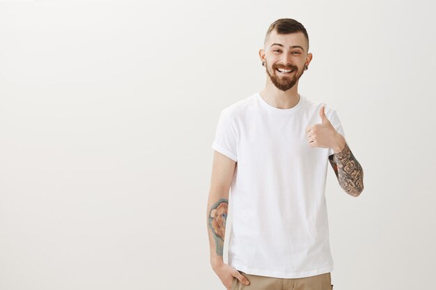 Hipster feliz satisfeito com tatuagens mostrando o polegar para cima, muito bem
