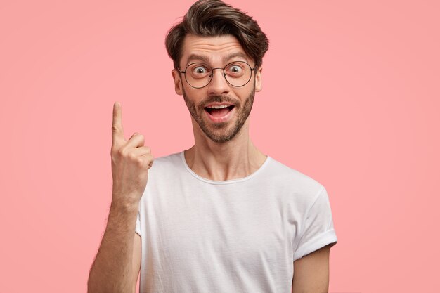 Hipster surpreso com corte de cabelo da moda, tem olhar intrigante e chocado, aponta com o dedo indicador para cima, usa camiseta branca casual e óculos, isolado sobre a parede rosa