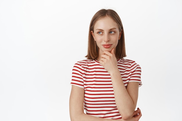 Hmm interessante. Jovem mulher bonita pensando, olhando para longe com um rosto sorridente e pensativo e toque no queixo, pondere a decisão, de pé na camiseta contra o fundo branco