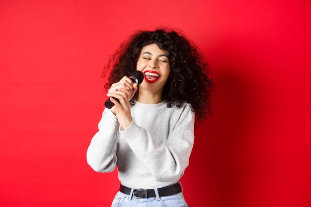 Hobbies e conceito de lazer. Mulher feliz cantando música no microfone, se divertindo no karaokê com microfone, em pé sobre fundo vermelho.