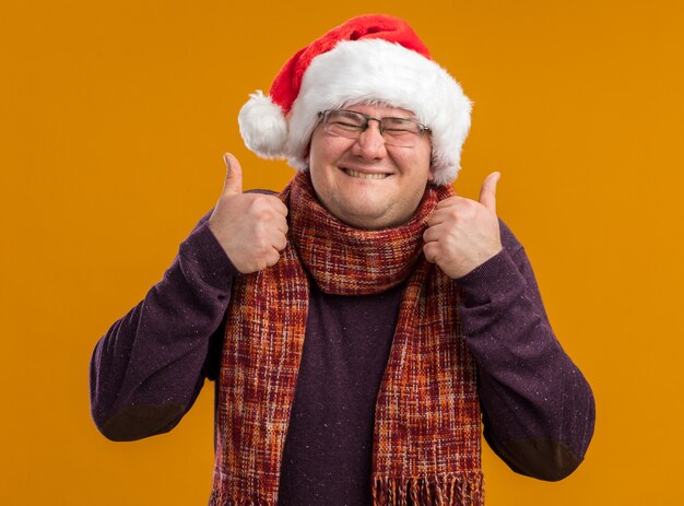 Homem adulto alegre usando óculos e chapéu de Papai Noel mordendo o lábio mostrando os polegares para cima com os olhos fechados, isolado em um fundo laranja