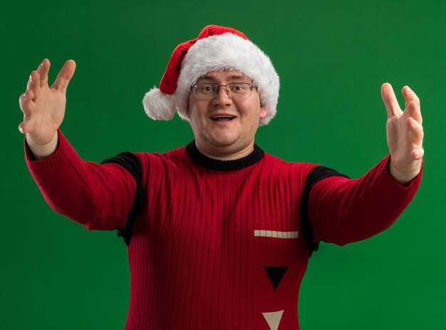 Foto grátis homem adulto amigável usando óculos e chapéu de papai noel olhando para a câmera fazendo gesto de boas-vindas isolado no fundo verde