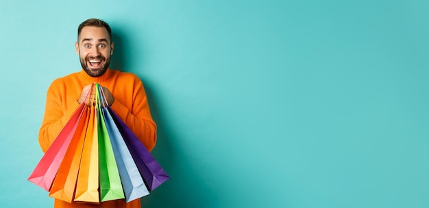 Foto grátis homem adulto animado segurando sacolas de compras e sorrindo indo ao shopping em pé sobre fundo turquesa