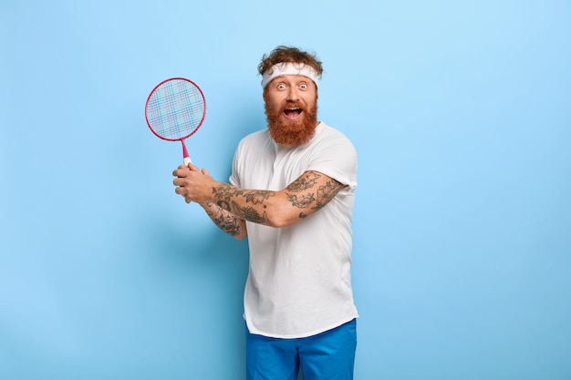 Homem adulto ativo e emotivo com cabelo ruivo segurando uma raquete de badminton e olhando feliz para a câmera