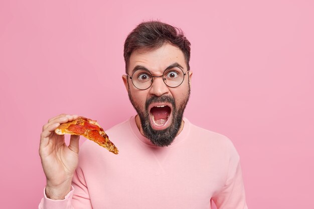 Homem adulto barbudo emocional gritando alto, segurando uma fatia de pizza saborosa e apetitosa, comendo fast food no lanche vestido com roupas casuais