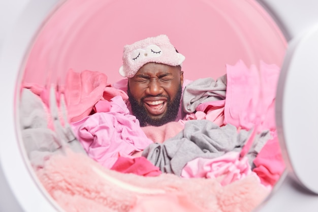 Homem adulto barbudo fecha os olhos e sorri amplamente usa máscara de dormir coberta com roupa lavada em casa posa contra a parede rosa