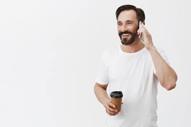 Homem adulto bonito e despreocupado tomando café e falando no smartphone