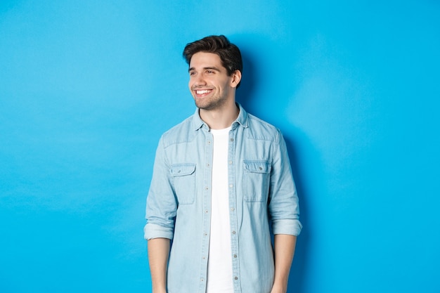 Homem adulto bonito e sorridente em roupa casual, sorrindo e olhando para a esquerda na oferta promocional, em pé contra um fundo azul