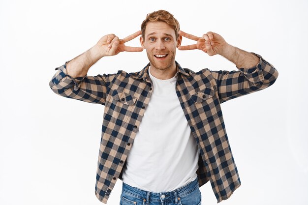 Homem adulto bonito engraçado dançando, mostrando o sinal V da paz e sorrindo, mantendo uma atitude positiva, animá-lo, brincar e mostrar rostos divertidos, em pé sobre uma parede branca