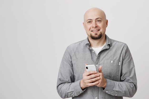 Homem adulto careca e sorridente feliz olhando de lado, segurando um smartphone