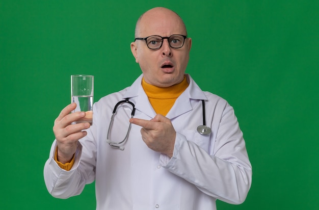 Foto grátis homem adulto chocado com óculos em uniforme de médico e estetoscópio segurando e apontando para um copo d'água