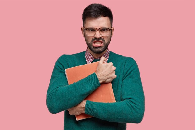 Homem adulto com barba por fazer irritado tem expressão irritada, mantém o livro com atenção, usa óculos com lentes grossas