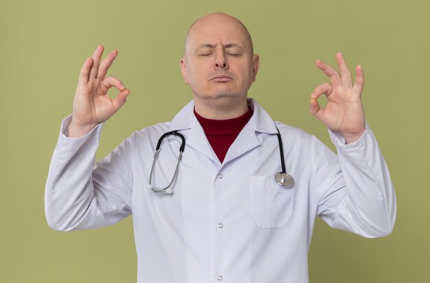 Homem adulto confiante em uniforme de médico com estetoscópio meditando em pé com os olhos fechados