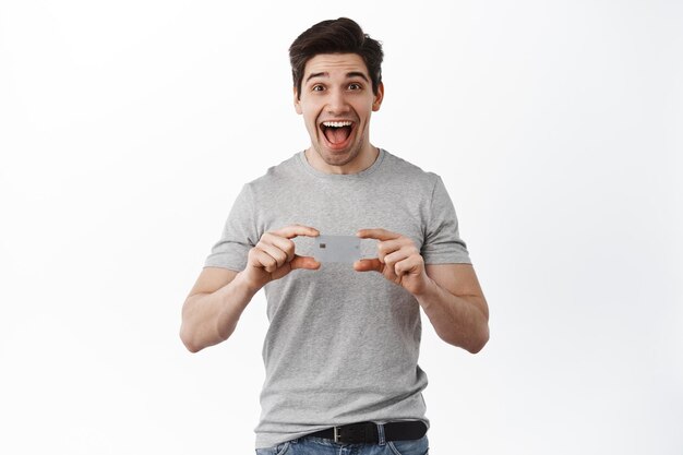 Foto grátis homem adulto empolgado mostrando cartão de crédito, gritando de espanto, mostrando fantástica promoção de banco, em pé sobre fundo branco