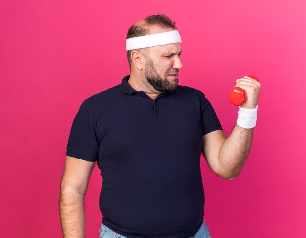 Homem adulto eslavo e desportivo desagradável usando bandana e pulseiras segurando e olhando para halteres isolado na parede rosa com espaço de cópia