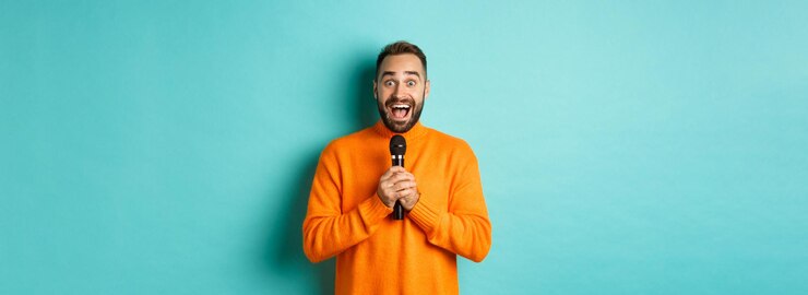 Foto grátis homem adulto feliz cantando karaokê segurando o microfone e olhando para a câmera em pé no suéter laranja