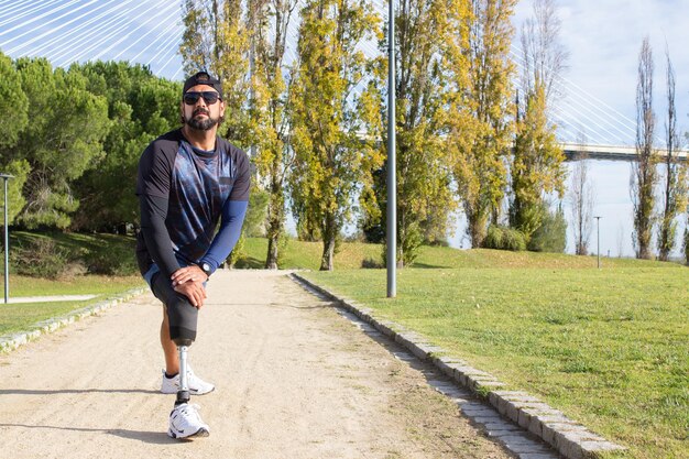 Homem adulto médio com perna artificial se preparando para correr. Homem em roupas esportivas esticando as pernas no parque em dia de verão. Esporte, conceito de treinamento