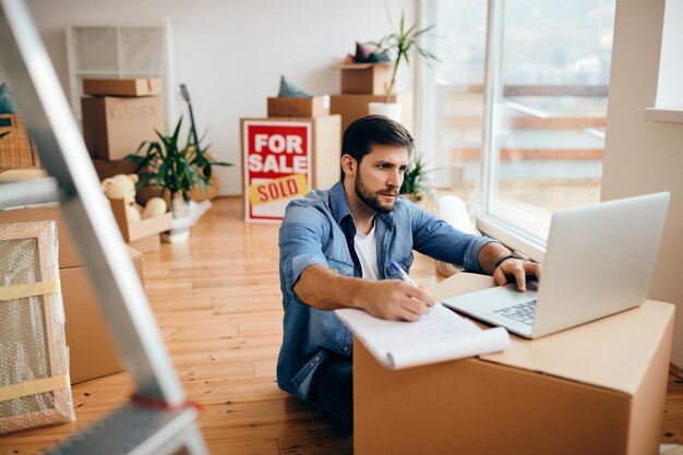 Homem adulto médio trabalhando em um computador e tomando notas em seu novo apartamento