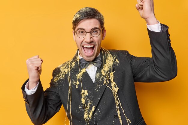 Foto grátis homem adulto positivo emocional vestido com roupas formais faz gesto de vencedor exclama em voz alta celebra sucesso manchado com spray serpantina isolado sobre fundo amarelo ocasião festiva