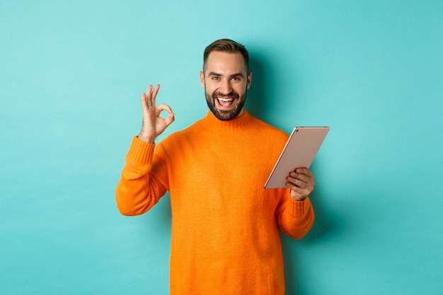 Homem adulto satisfeito sorrindo, usando tablet digital e mostrando sinal de ok, aprovo e concordo, em pé