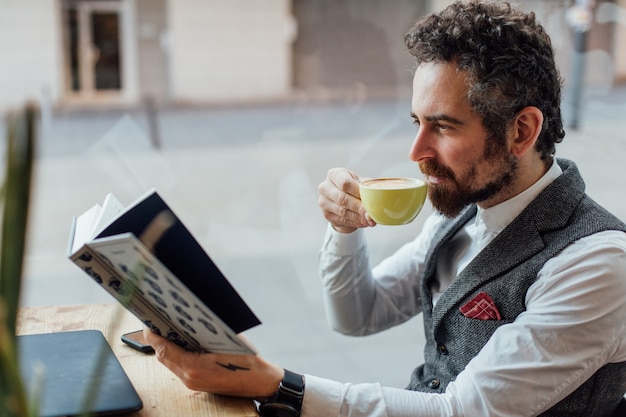 Homem adulto sério de meia-idade bebe e aprecia uma bebida de café enquanto lê um livro interessante e cativante em um café ou biblioteca
